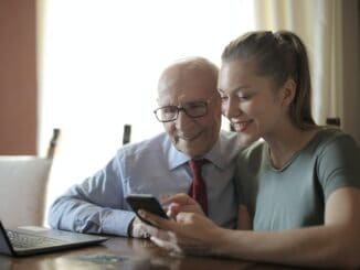 Es sind 2 Personen zu sehen. Ein älterer Mann in Hemd und Krawatte und eine junge Frau. Sie lächeln beide und schauen in ein Smartphone. Sie sitzen nebeneinander und vor ihnen steht ein Laptop.