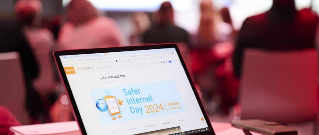 Ein geöffneter Laptop mit dem Logo des Safer-Internet-Days am Bildschirm. Im Hintergrund sind Menschen und eine Bühne zu sehen. ©Saferinternet.at