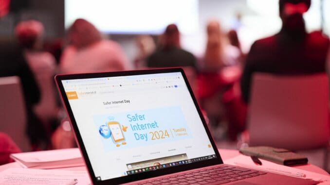 Ein geöffneter Laptop mit dem Logo des Safer-Internet-Days am Bildschirm. Im Hintergrund sind Menschen und eine Bühne zu sehen. ©Saferinternet.at