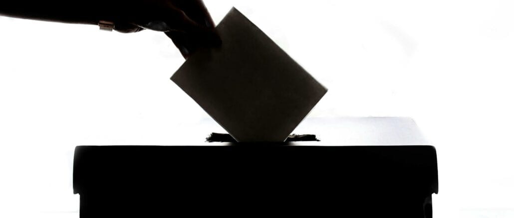 Zu sehen ist eine Hand, die einen Umschlag in eine Wahlurne steckt.