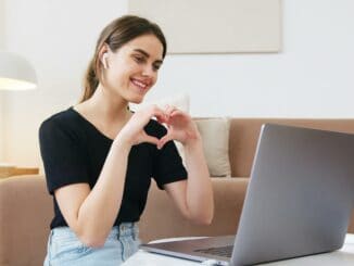 Zu sehen ist eine Frau, die ein Herz mit ihren Händen formt. Sie nimmt aktuell an einem Video-Call teil. Sie sitzt vor ihrem Laptop.