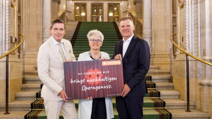 Zu sehen sind drei Personen auf der Feststoege der Wiener Staatsoper. Die Dame in der Mitte hält ein Schild hoch, auf welchem zu lesen ist: 