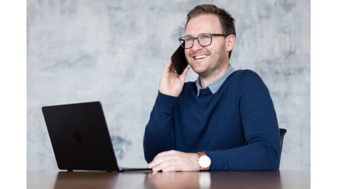 Zu sehen ist Daniel Gaubinger-Börner, Barrierefreiheitsexperte. Er sitz an einem Tisch mit seinem Laptop und telefoniert dabei.