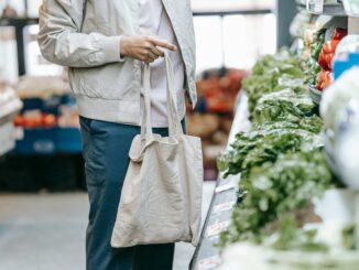 Zu sehen ist ein Mensch mit Einkaufstausche. Der Kopf ist nicht zu sehen. Die Person steht in einem Supermarkt und Gemüse ist vor der Person aufgeschichtet.