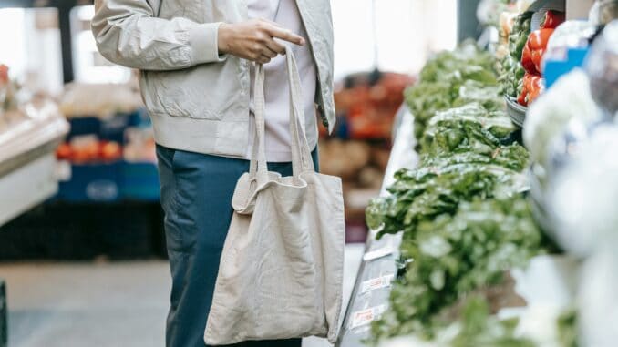 Zu sehen ist ein Mensch mit Einkaufstausche. Der Kopf ist nicht zu sehen. Die Person steht in einem Supermarkt und Gemüse ist vor der Person aufgeschichtet.