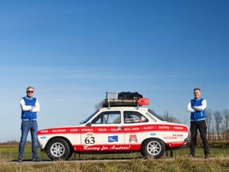 Zu sehen sind Müller und Huber auf einem Feldweg. Zwischen den beiden Männern mit verschränkten Armen steht der Ford Escort.